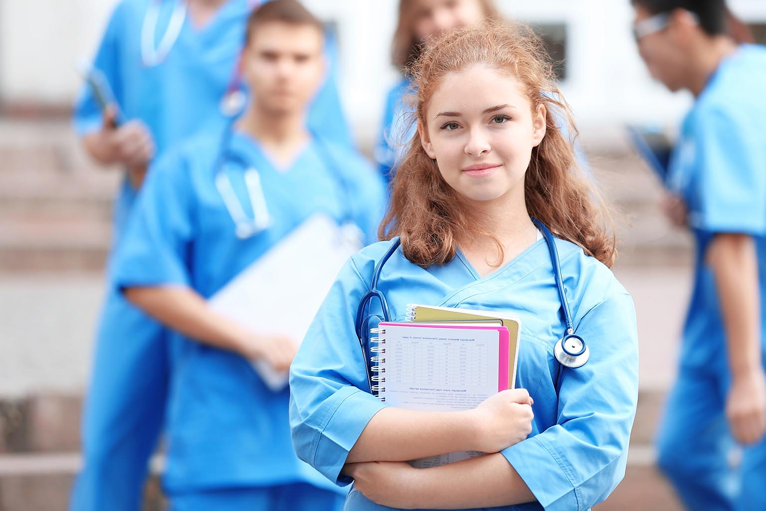 Nursing student in blue scrubs holding books with classmates on a campus staircase